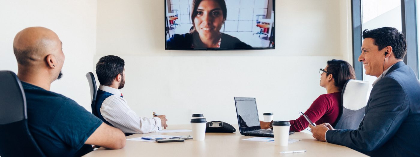 A team around a conference table with one colleague joining on screen, seen from the end of the room.